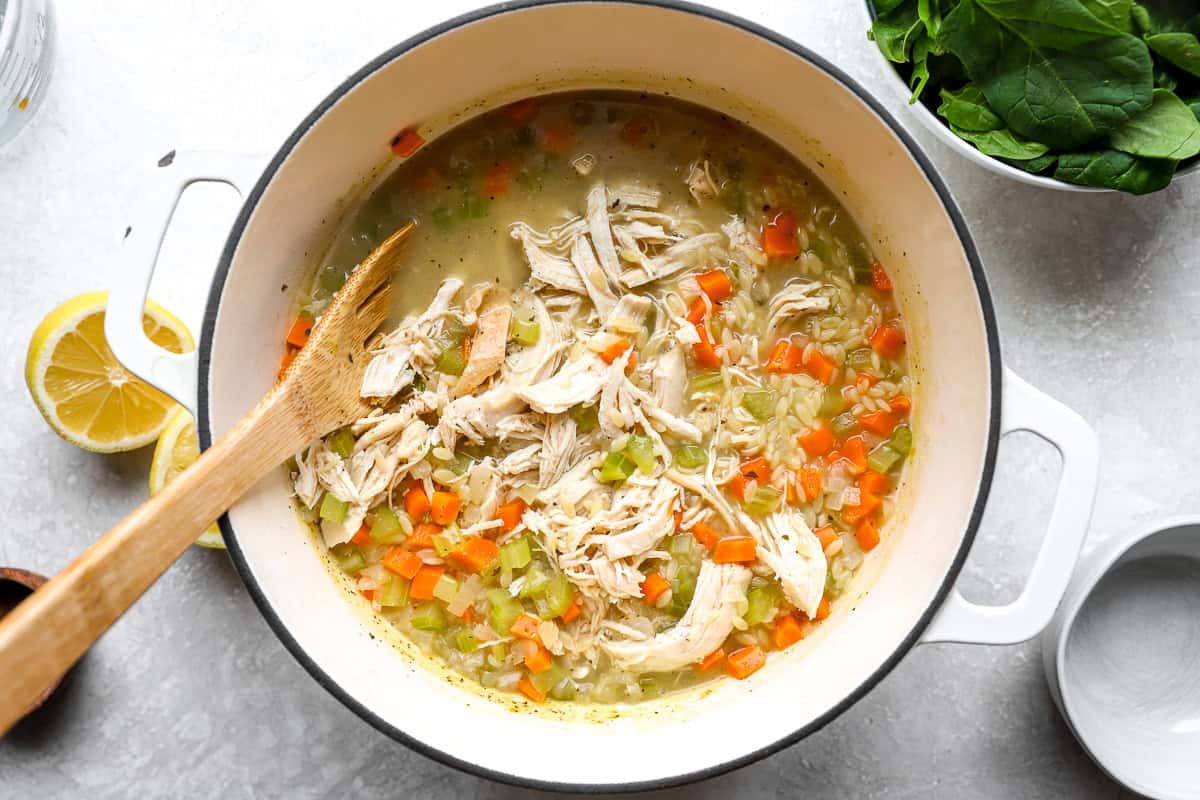 Shredded chicken being added to lemon chicken orzo soup with vegetables in a large pot