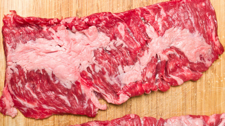A piece of raw skirt steak with big globs of fat sitting on a cutting board.