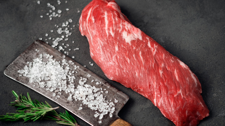 A piece of skirt steak on a marbled black table with rock salt and rosemary sprigs.