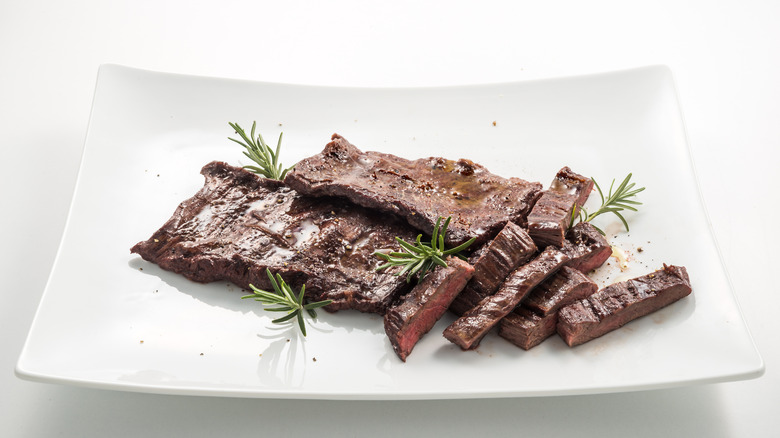 Half-sliced skirt steak on a white. curved, rectangular plate with sprigs of rosemary.