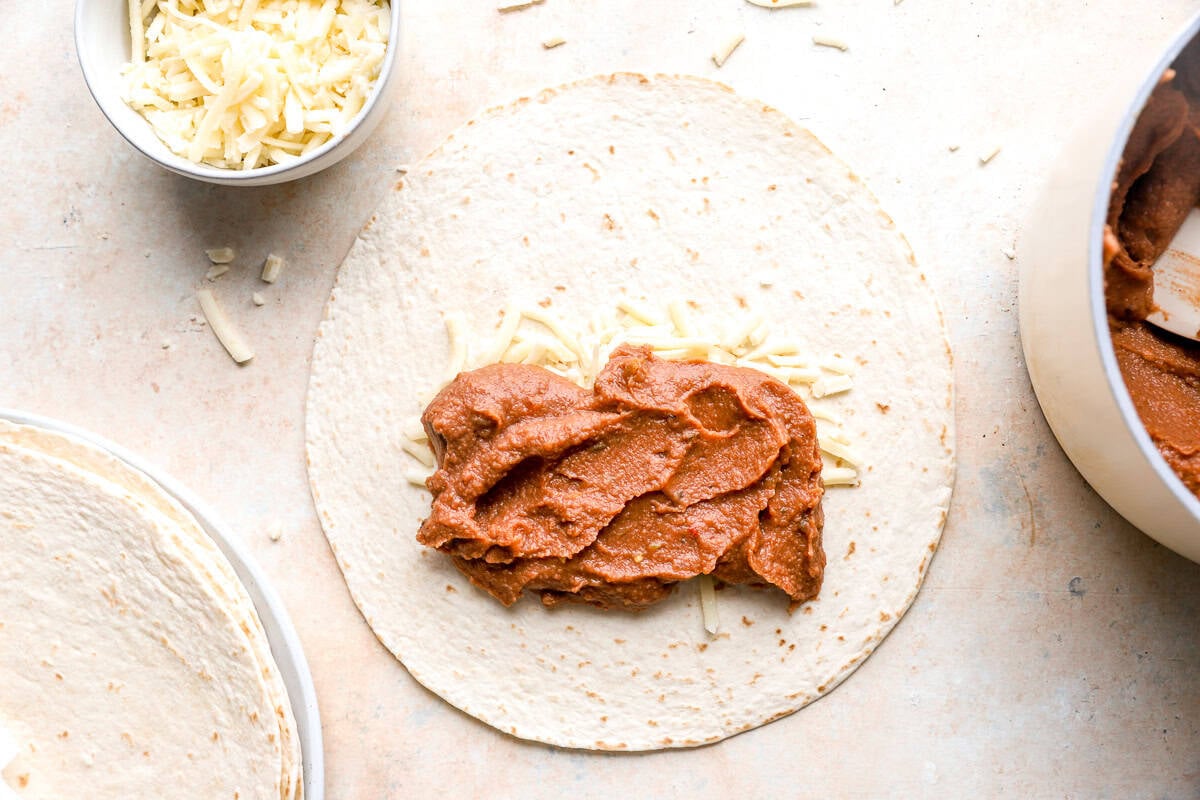 Refried beans and shredded cheese layered onto a flour tortilla before rolling.