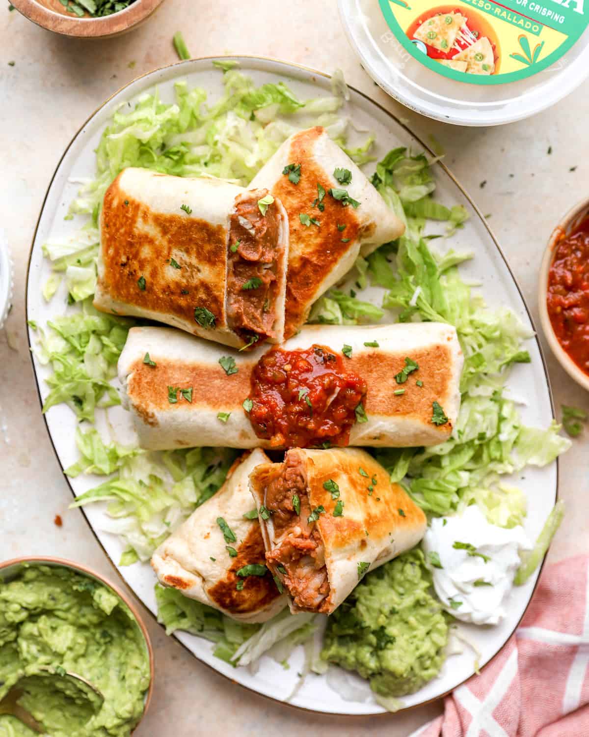 Crispy bean and cheese burritos served with guacamole, salsa, sour cream, and shredded lettuce on a platter.