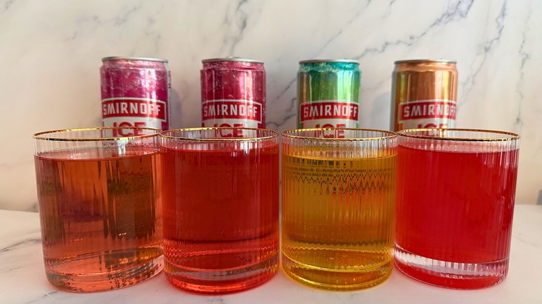 Four glasses of colored beverage in front of four cans of Smirnoff Ice