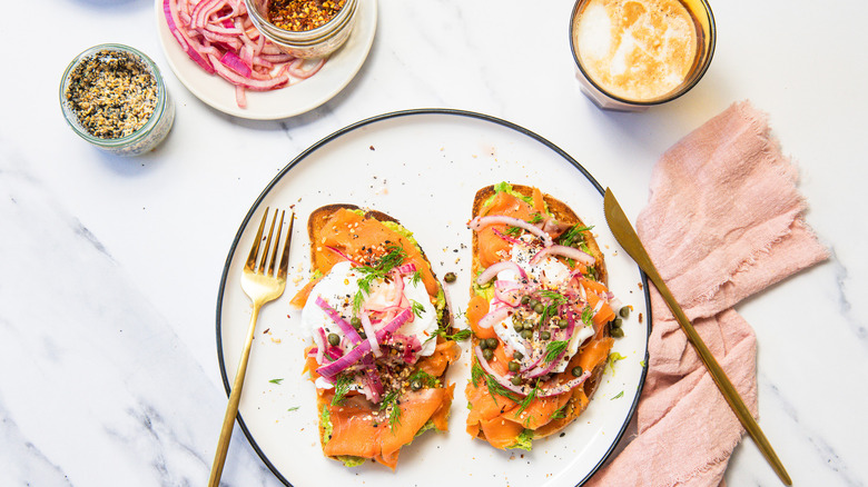 Two slices of smoked salmon breakfast toast on white plate with coffee, cutlery and napkin.