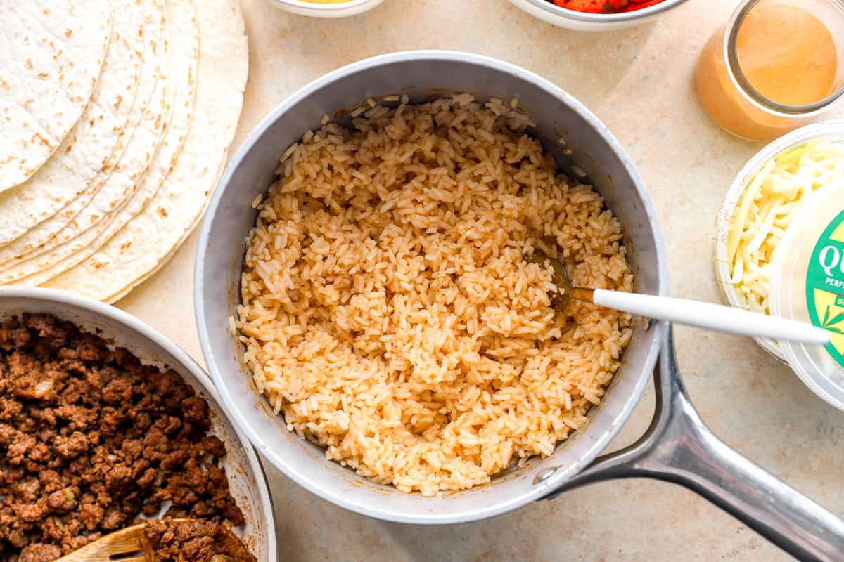 Taco-seasoned rice in a saucepan surrounded by tortillas, cheese, and burrito ingredients.