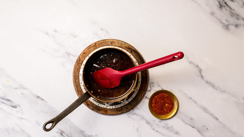 Hot honey in saucepan with spatula beside pinch bowl with ground Thai chile flakes.