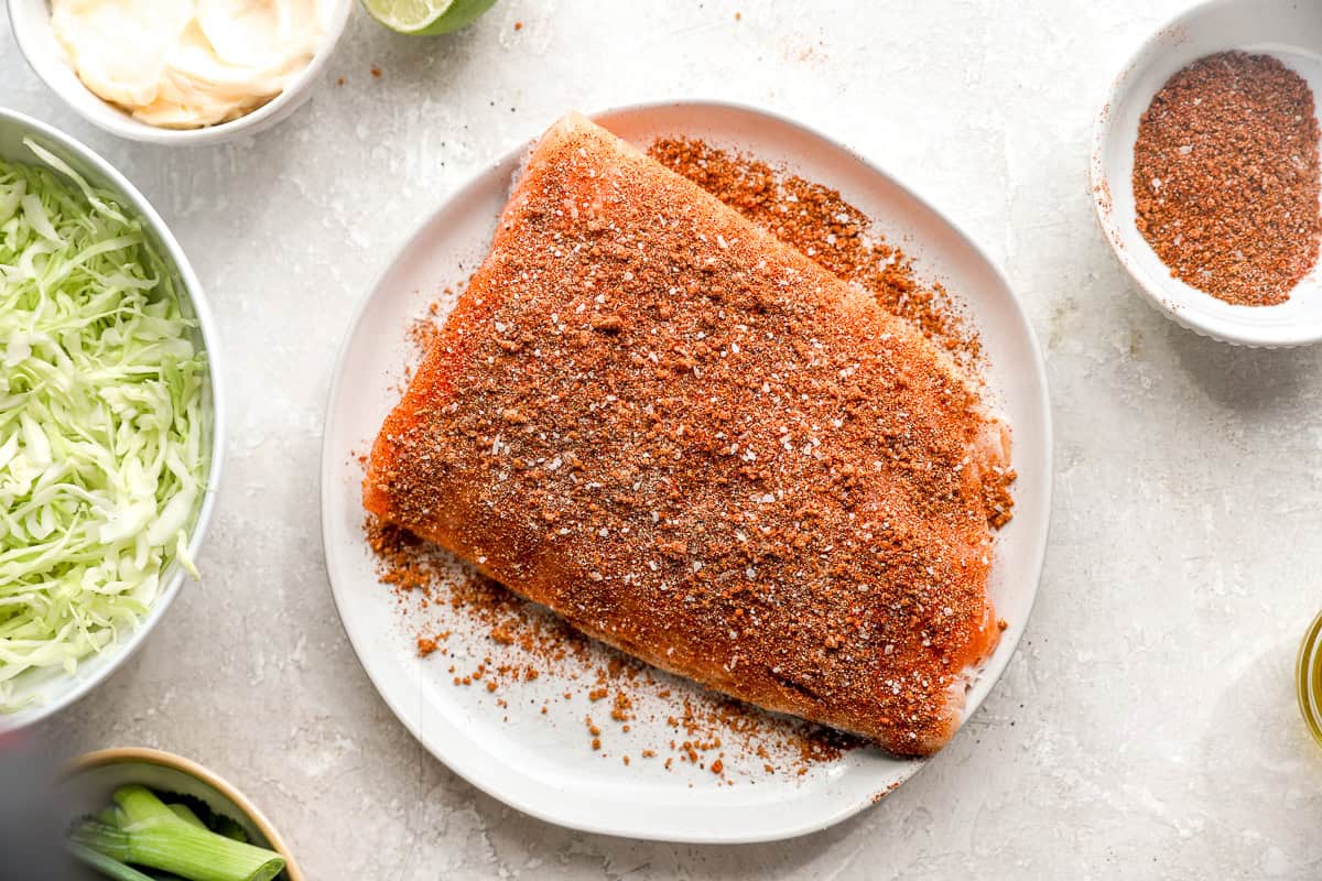 Salmon filet coated in smoky taco seasoning on a plate surrounded by cabbage, lime, and taco ingredients.