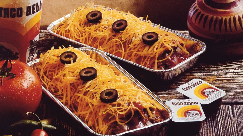Two Taco Bell enchiritos  in foil containers, covered in cheese and olives  with a drink and taco sauce to the sides