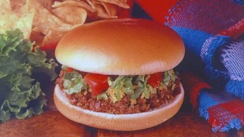 A Taco Bell chili burger sandwich featuring meat, tomato, and lettuce on a hamburger bun with tortilla chips and a blanket in the background
