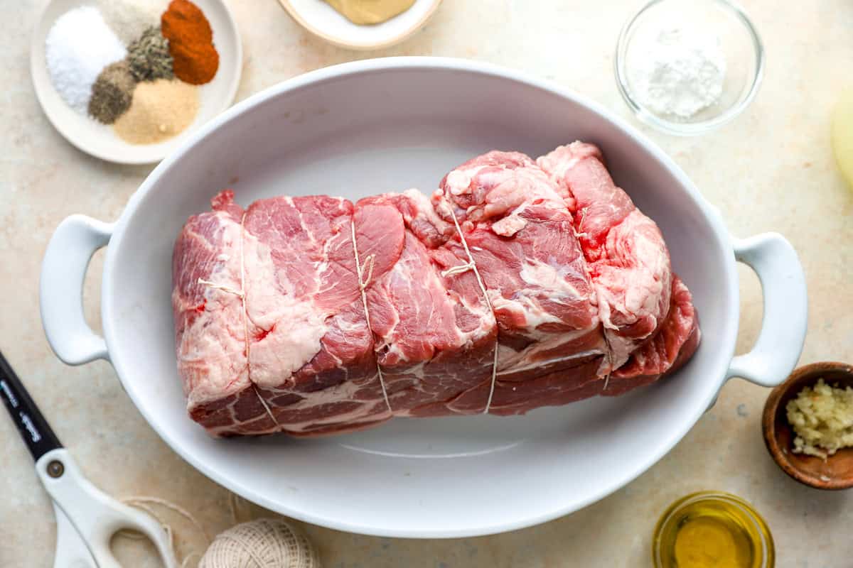 Raw pork shoulder roast tied with kitchen twine in a white baking dish with seasonings nearby.