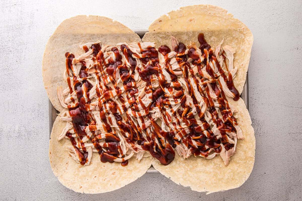 Shredded rotisserie chicken layered over tortillas and drizzled with BBQ sauce on a sheet pan.