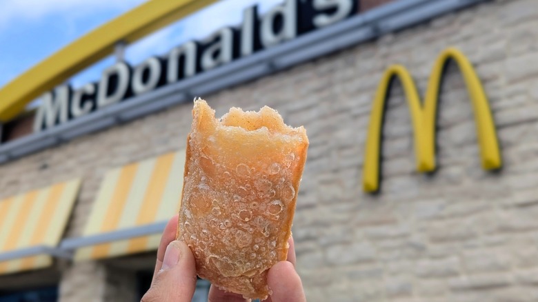 A hand holding a half-eaten Fired Apple Pie in front of the McDonald's storefront