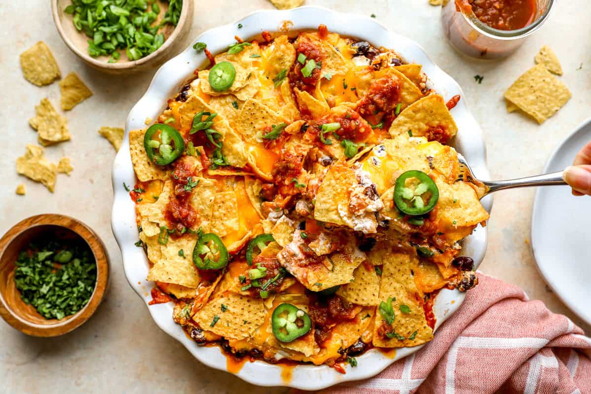 Spoon lifting a cheesy serving of nacho pie topped with salsa, jalapeños, and green onions from the baking dish