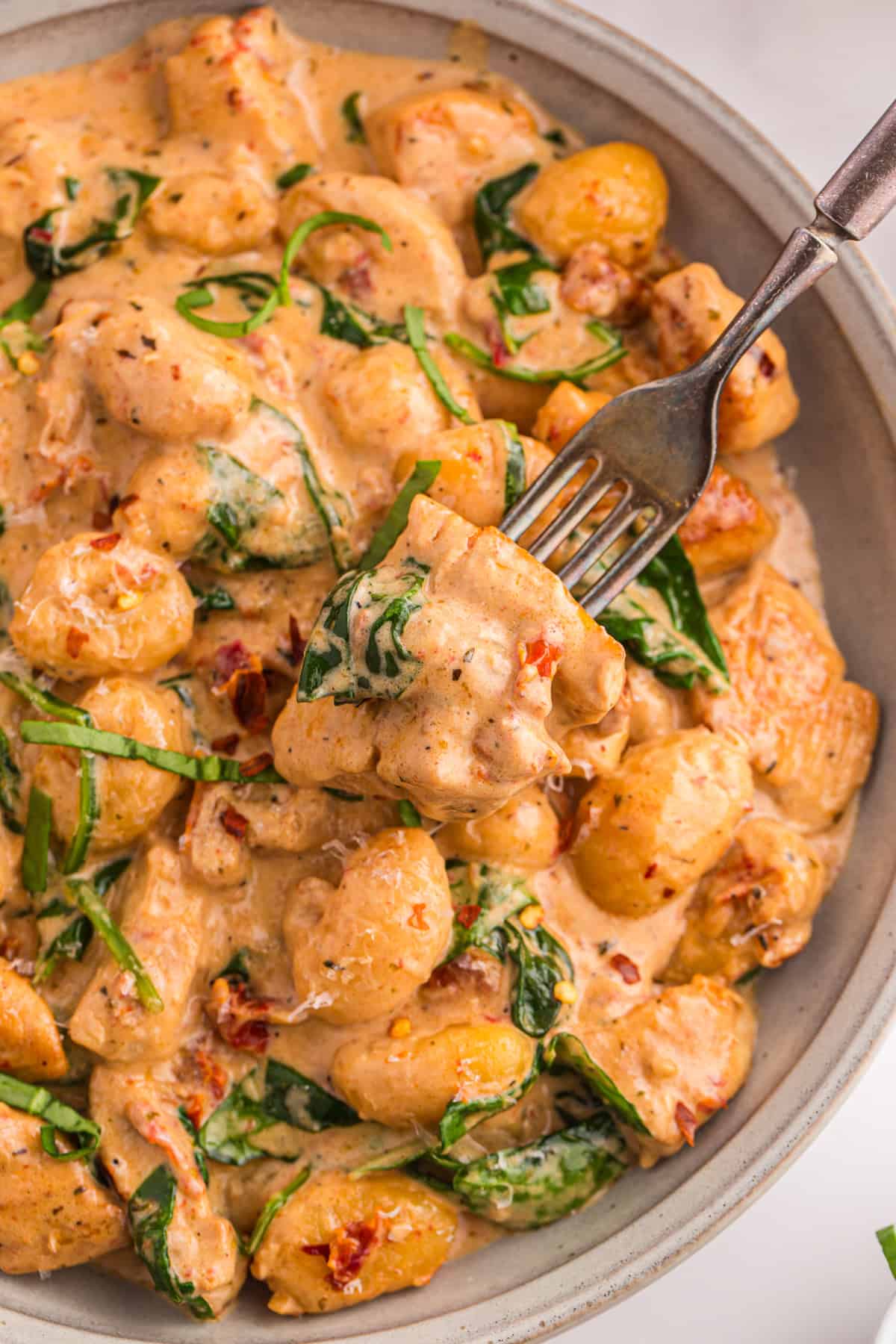 Overhead close-up of marry me chicken gnocchi in a bowl with a fork lifting a piece of chicken, showing the creamy sun-dried tomato sauce and wilted spinach.