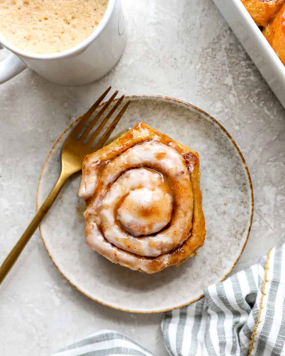 Heavy cream cinnamon roll served on a plate with cream cheese icing and coffee in the background.