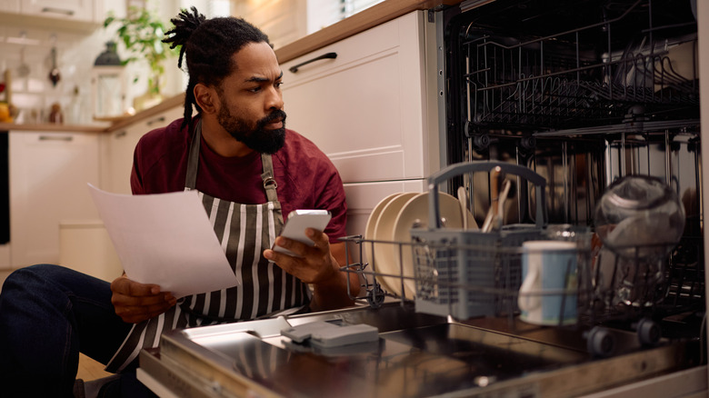 A man using his phone and a dishwasher manual to determine how to use the appliance