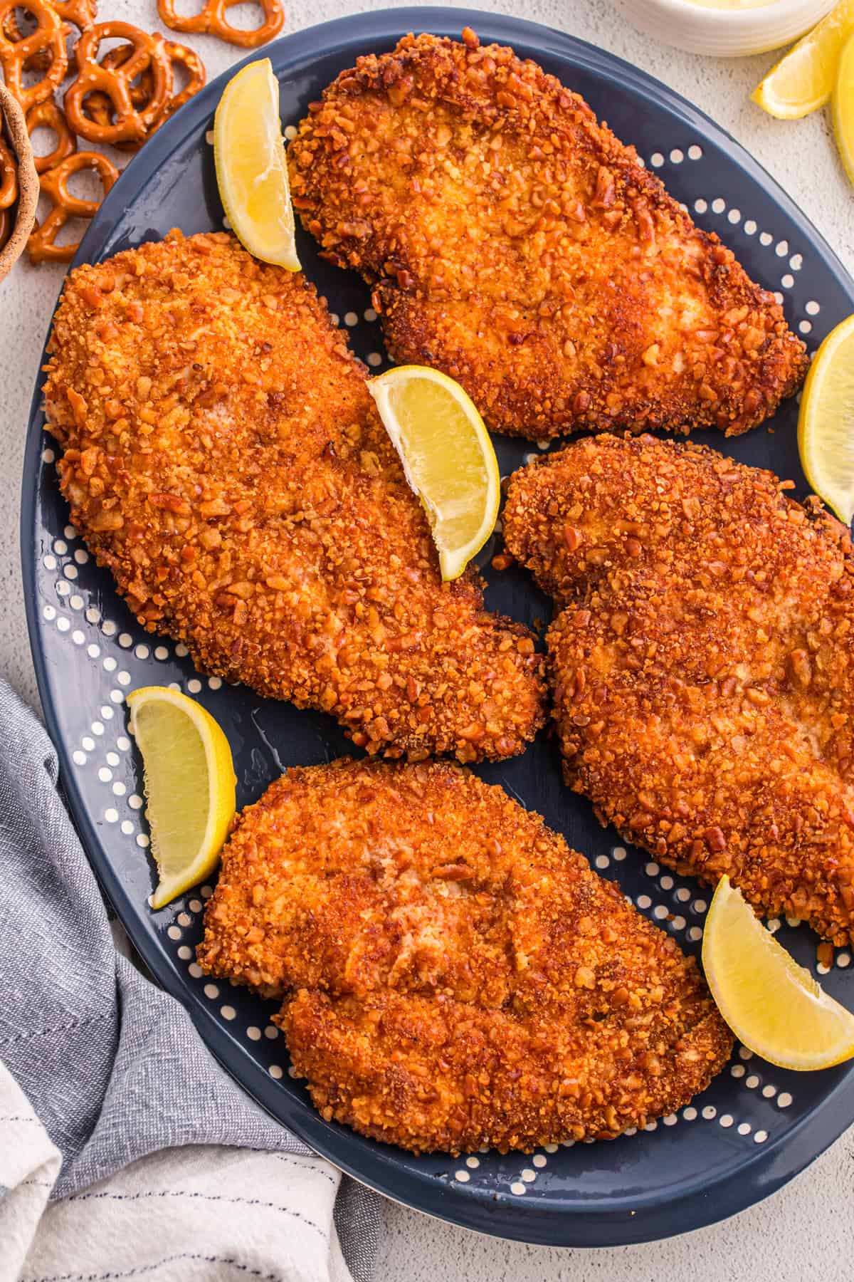 Pretzel crusted chicken cutlets on a dark blue oval platter with lemon wedges and whole pretzels in the background