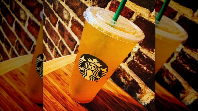A Starbucks Orange Vanencia Refresh in a branded plastic cup sits on a wooden table, with an exposed-brick wall in the background