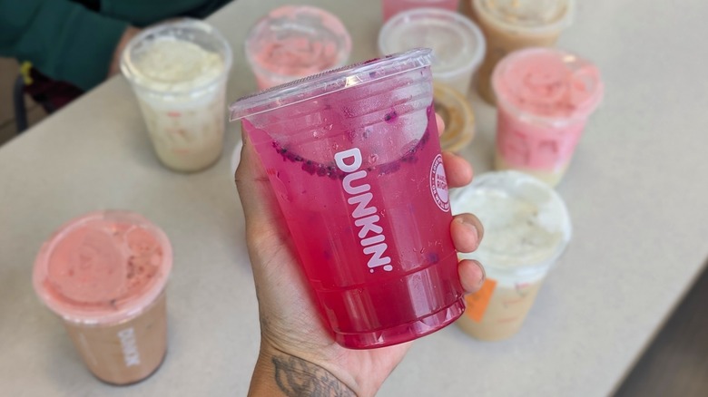 A hand holding a bright pink Dunkin' drink with more Dunkin' drinks on a table in the background