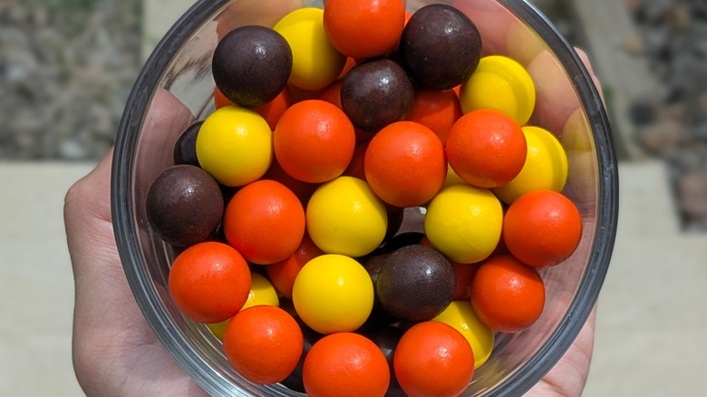 A close-up of a hand holding round orange, brown, and yellow candies in a glass bowl