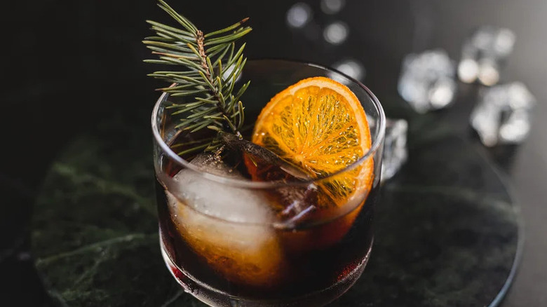 Close-up of dark cocktail with rosemary sprig and orange wheel garnish against black background.