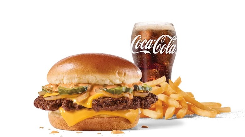 A classic Jack in the Box cheeseburger, fries and Coca-Cola in logo glass on white background