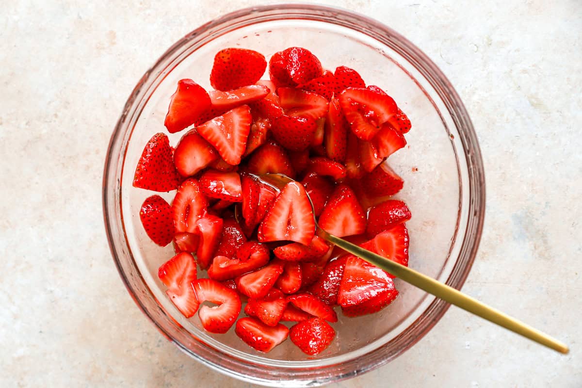 Sliced strawberries macerating in sugar in a glass bowl with a gold spoon