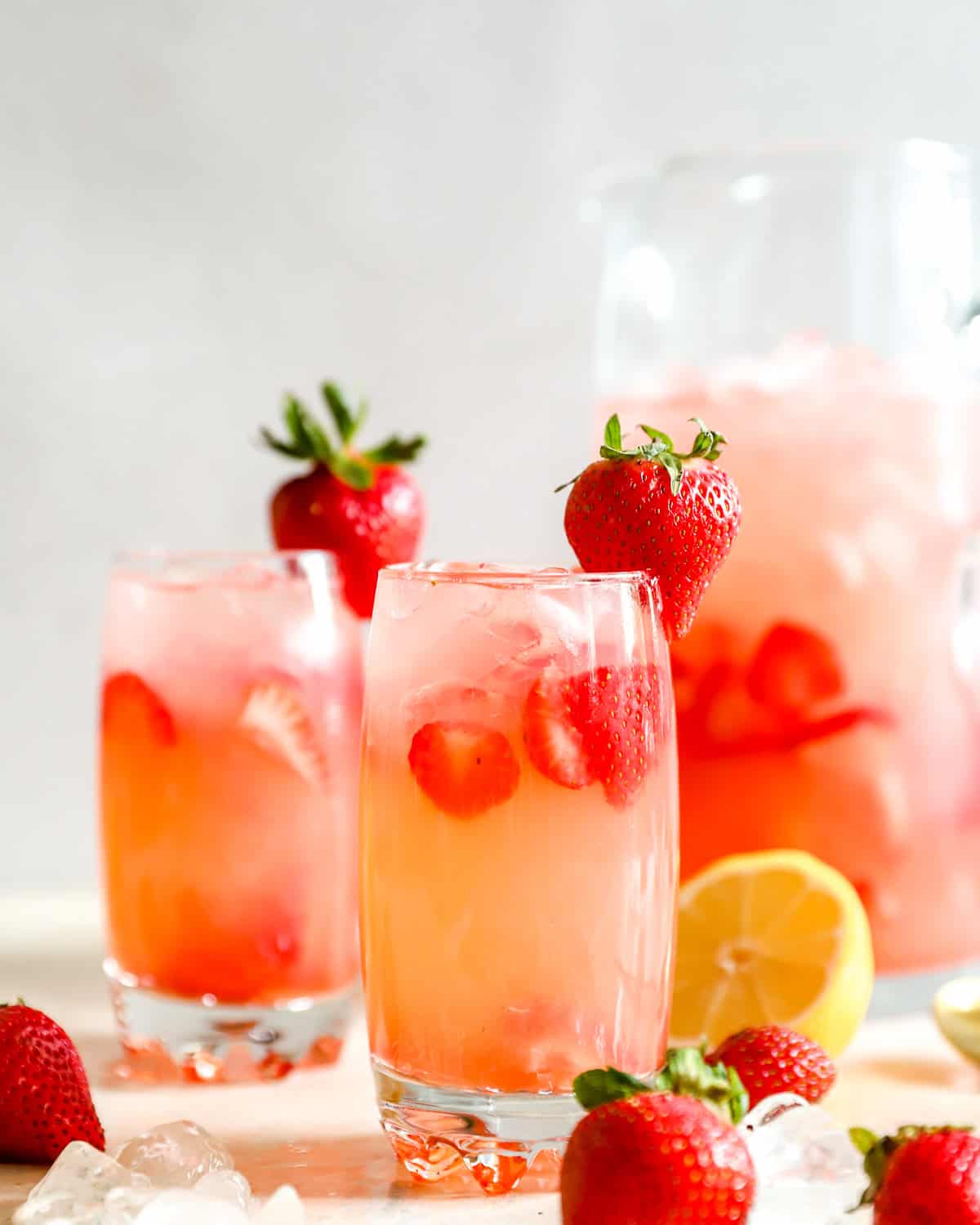 Two glasses of copycat Red Robin strawberry lemonade with sliced strawberries and ice, with a whole strawberry garnish and a pitcher behind them