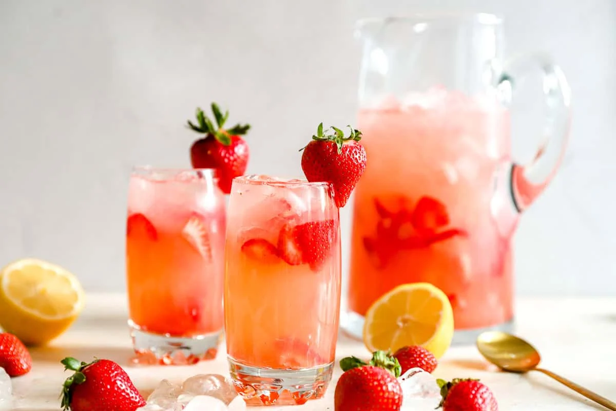 Pitcher and two glasses of homemade strawberry lemonade with fresh strawberries, lemon halves, and ice scattered around