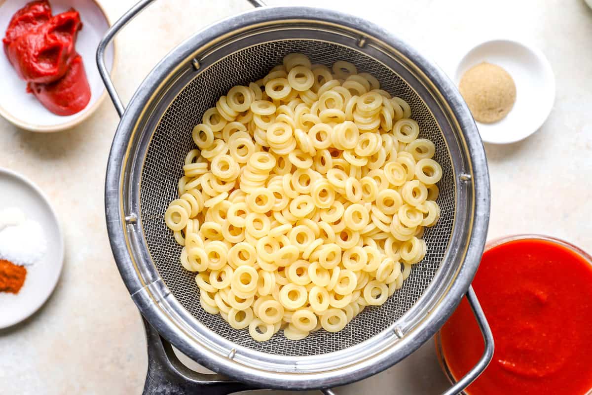 Cooked anellini pasta draining in a mesh strainer, with tomato paste, brown sugar, and tomato sauce in bowls nearby.