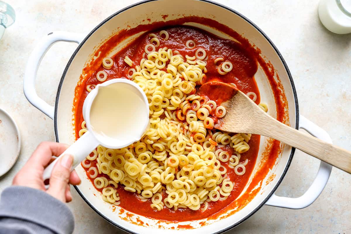 Pouring reserved pasta water into the pot as cooked anellini pasta is stirred into the tomato sauce.