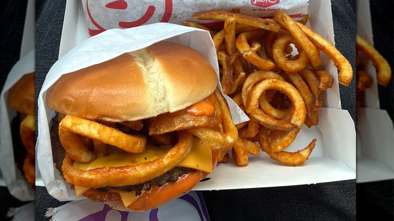 Jack In The Box burger topped with visible curly fries sitting on paper next to it