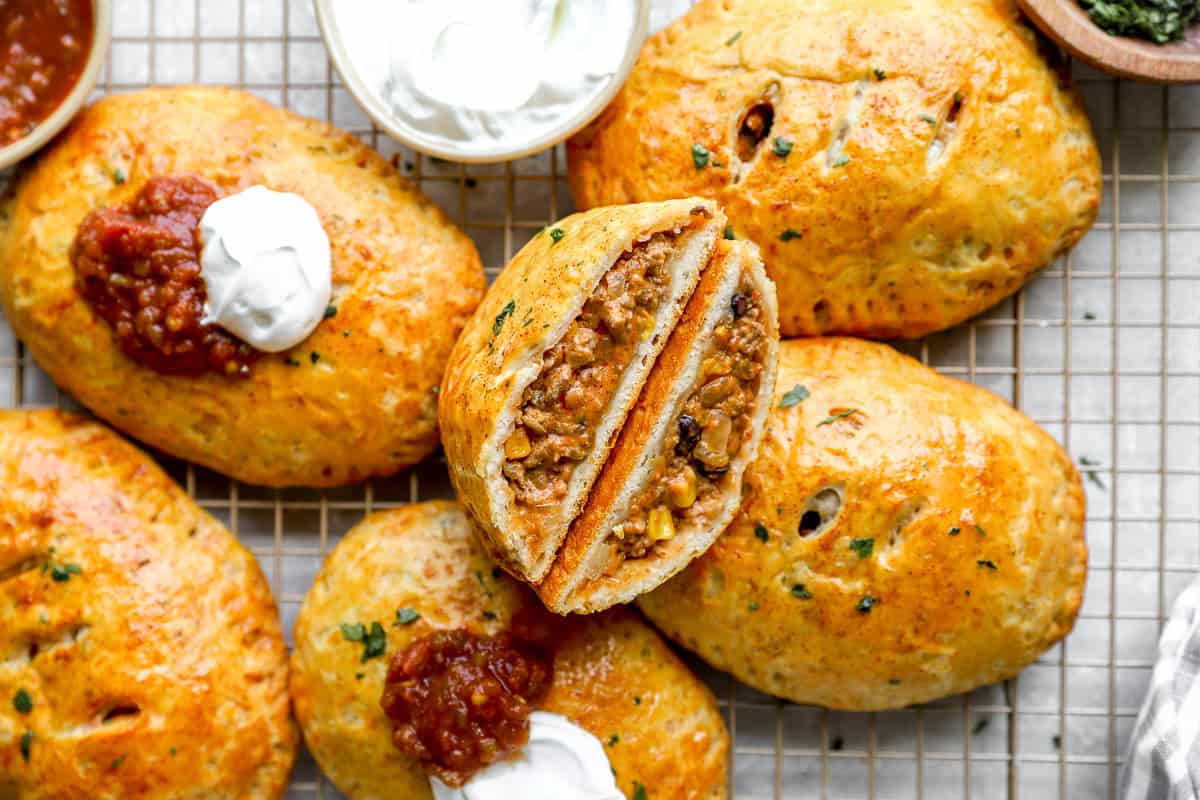 Baked Loaded Taco Hot Pockets on a wire cooling rack, topped with salsa and sour cream and one sliced open.