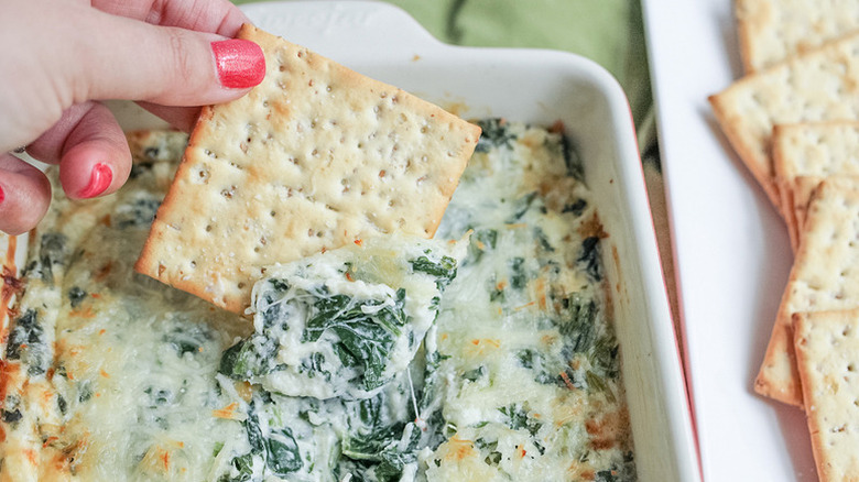Hand dipping cracker into dish of creamy spinach dip