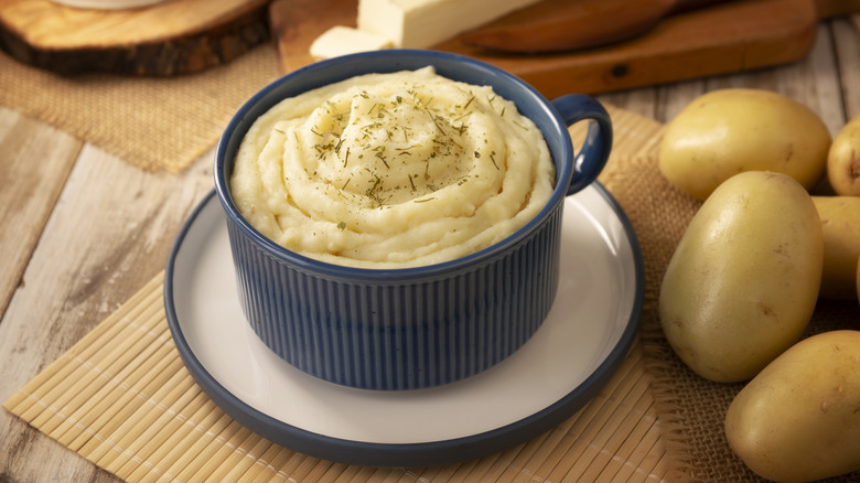 Crock of mashed potatoes on a wooden board next to some whole potatoes