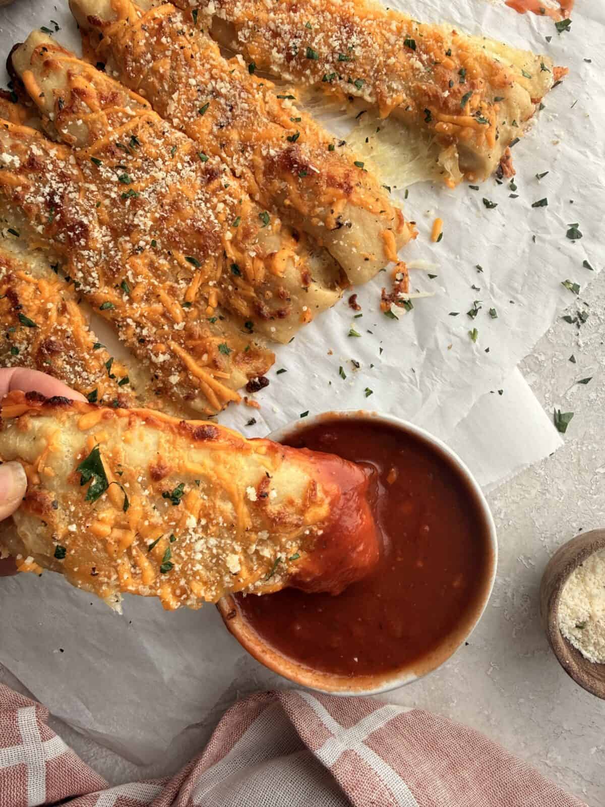 Hand dipping a cheesy breadstick into a bowl of marinara sauce, showing the melted cheese pull