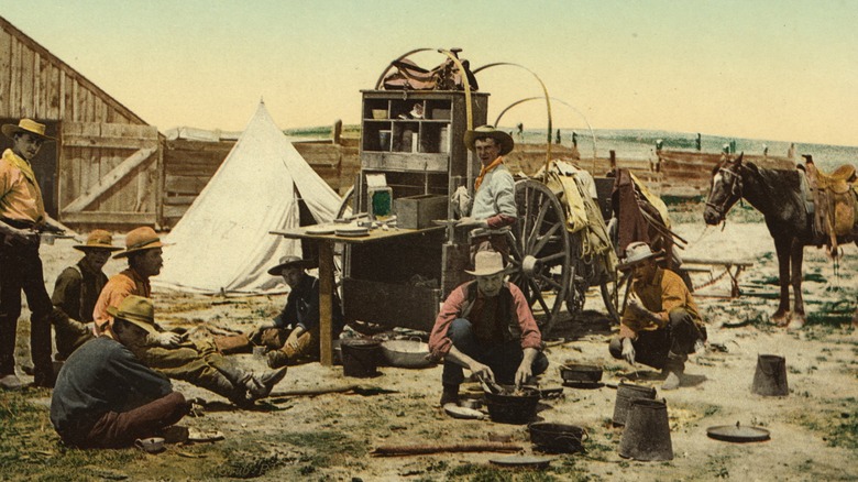 A photo of cowboys sharing a meal with their belongings, a tent, a makeshift table top, and a horse with a saddle spread throughout the scene