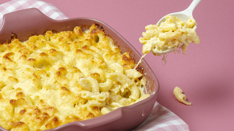 a spoon of cheesy macaroni lifted from a tray of baked mac and cheese on pink background