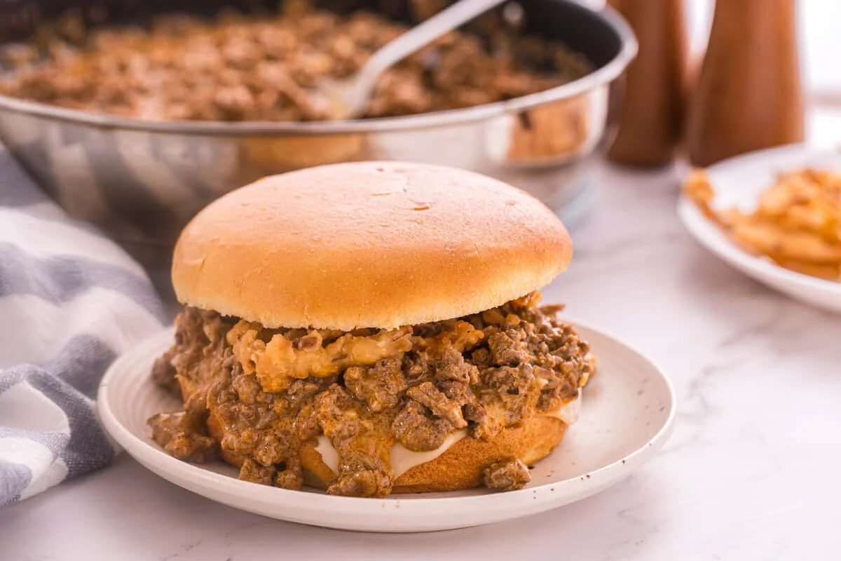 Front view of an assembled Beef Stroganoff Sloppy Joe sandwich on a plate with the skillet of filling and crispy fries in the background