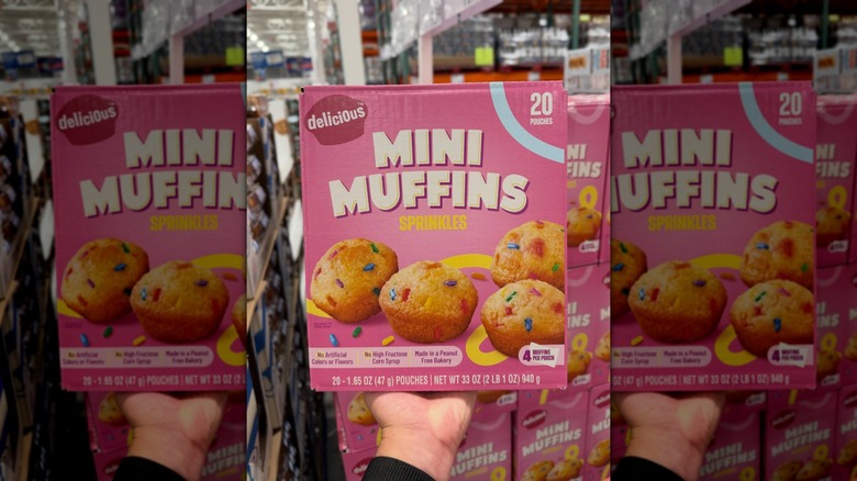 A woman's hand holding up a pink box of Delicious Mini Muffins Sprinkles in a Costco warehouse