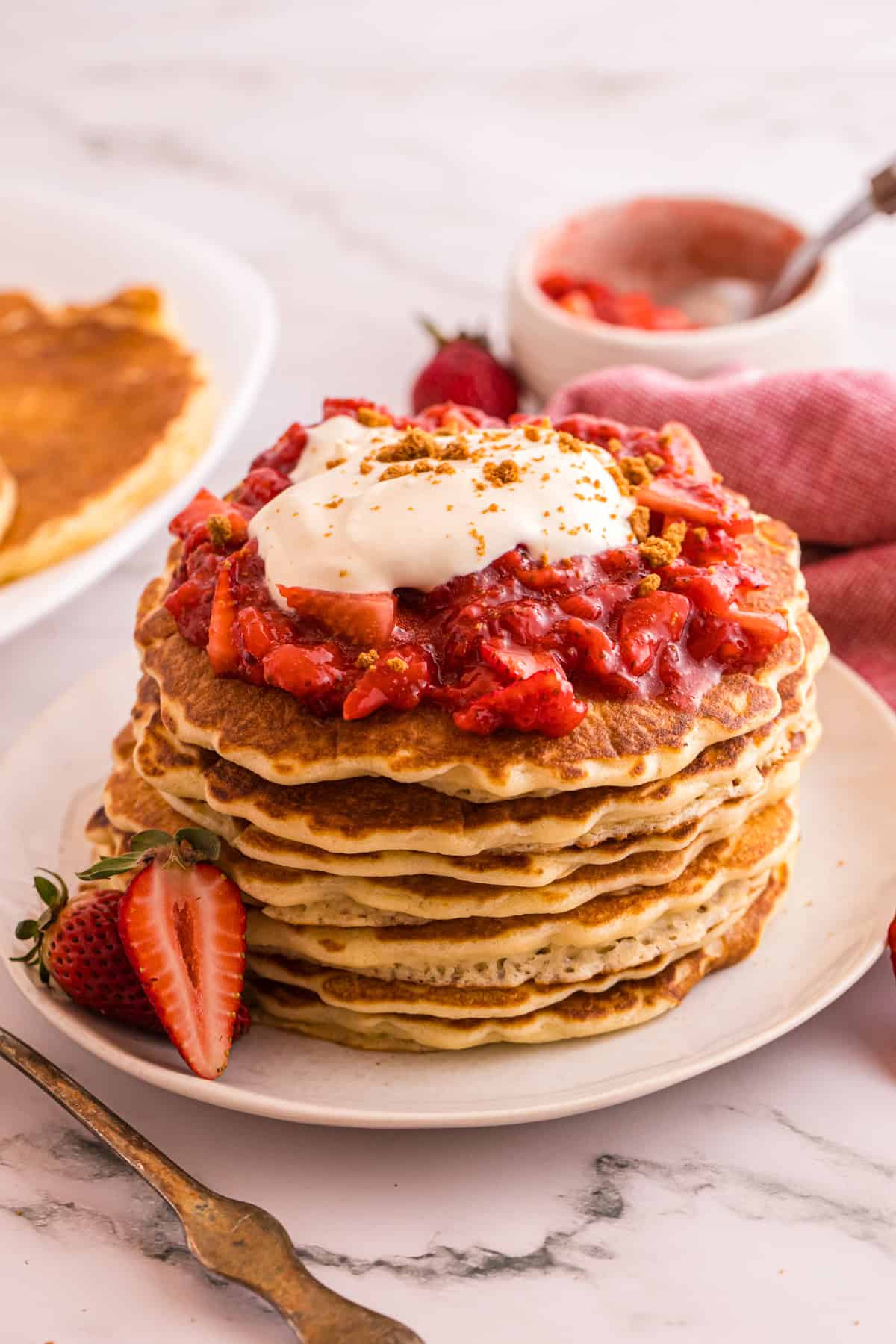Stack of strawberry cheesecake pancakes topped with strawberry sauce, whipped cream, and graham cracker crumbs on a white plate