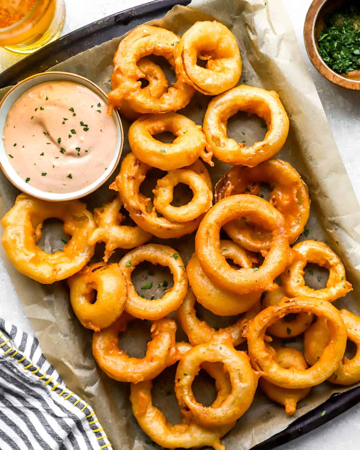 Overhead shot of crispy homemade onion rings piled on a parchment-lined tray with a bowl of pink dipping sauce, a bowl of chopped herbs, and a glass of beer alongside.