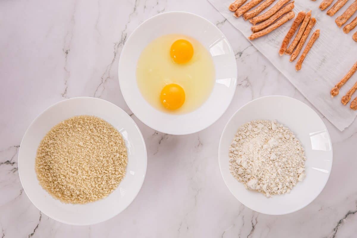 Overhead view of a breading station with panko breadcrumbs, beaten eggs, and flour in separate bowls, next to raw chicken strips.