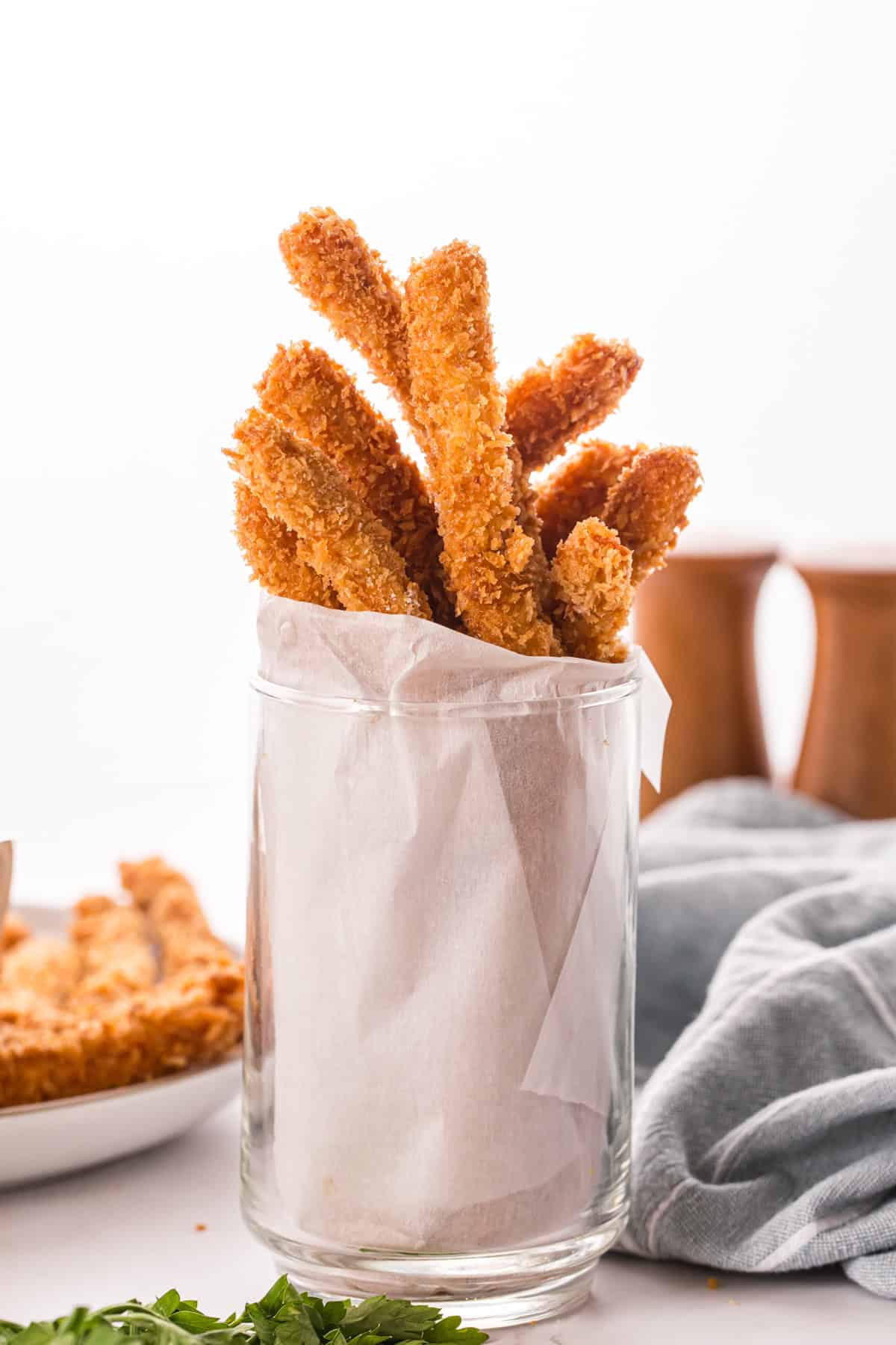 Crispy breaded chicken fries standing upright in a parchment-lined glass, with more fries and dipping sauce blurred in the background.