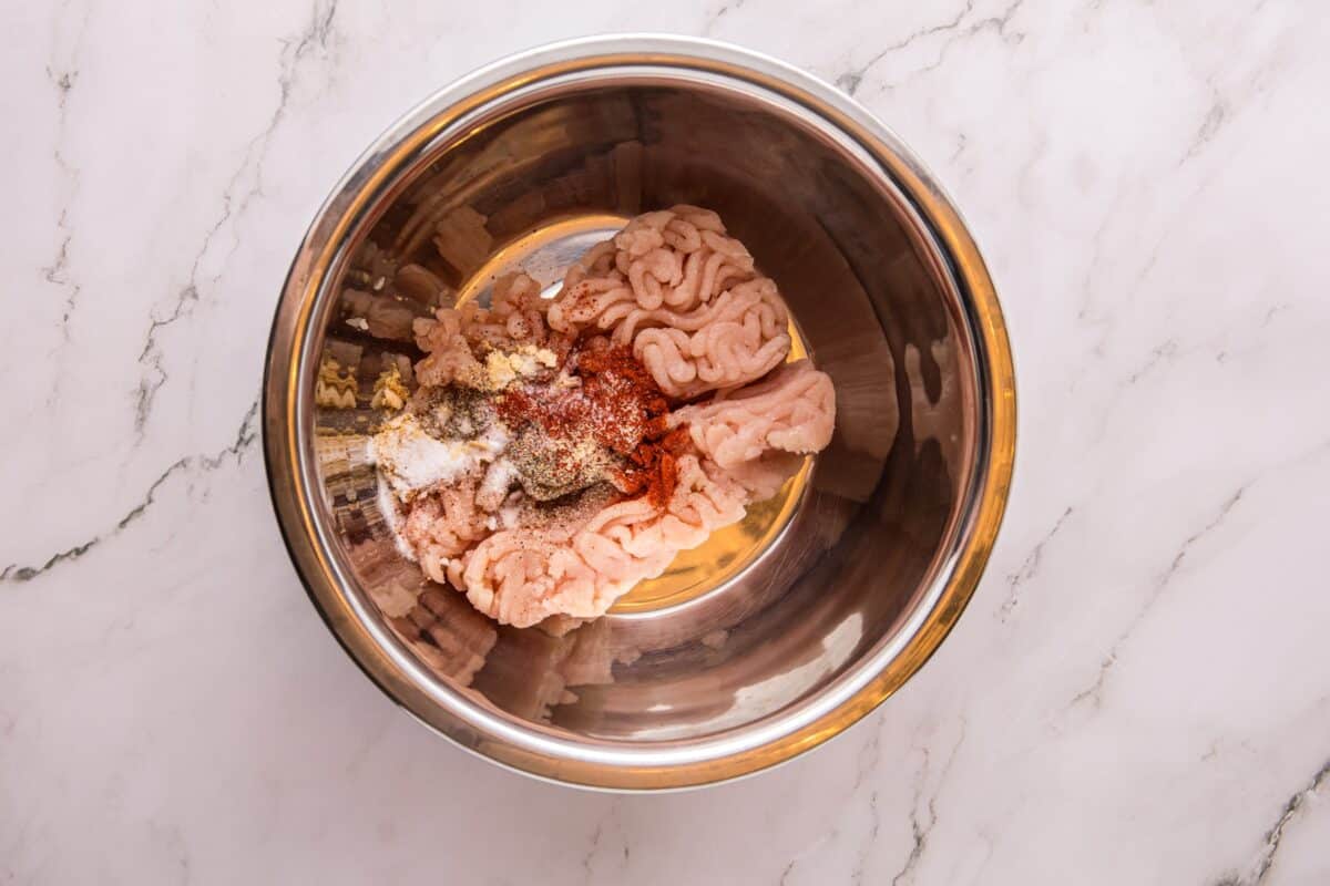 Overhead view of ground chicken and seasonings, including paprika, garlic powder, and black pepper, in a metal mixing bowl.