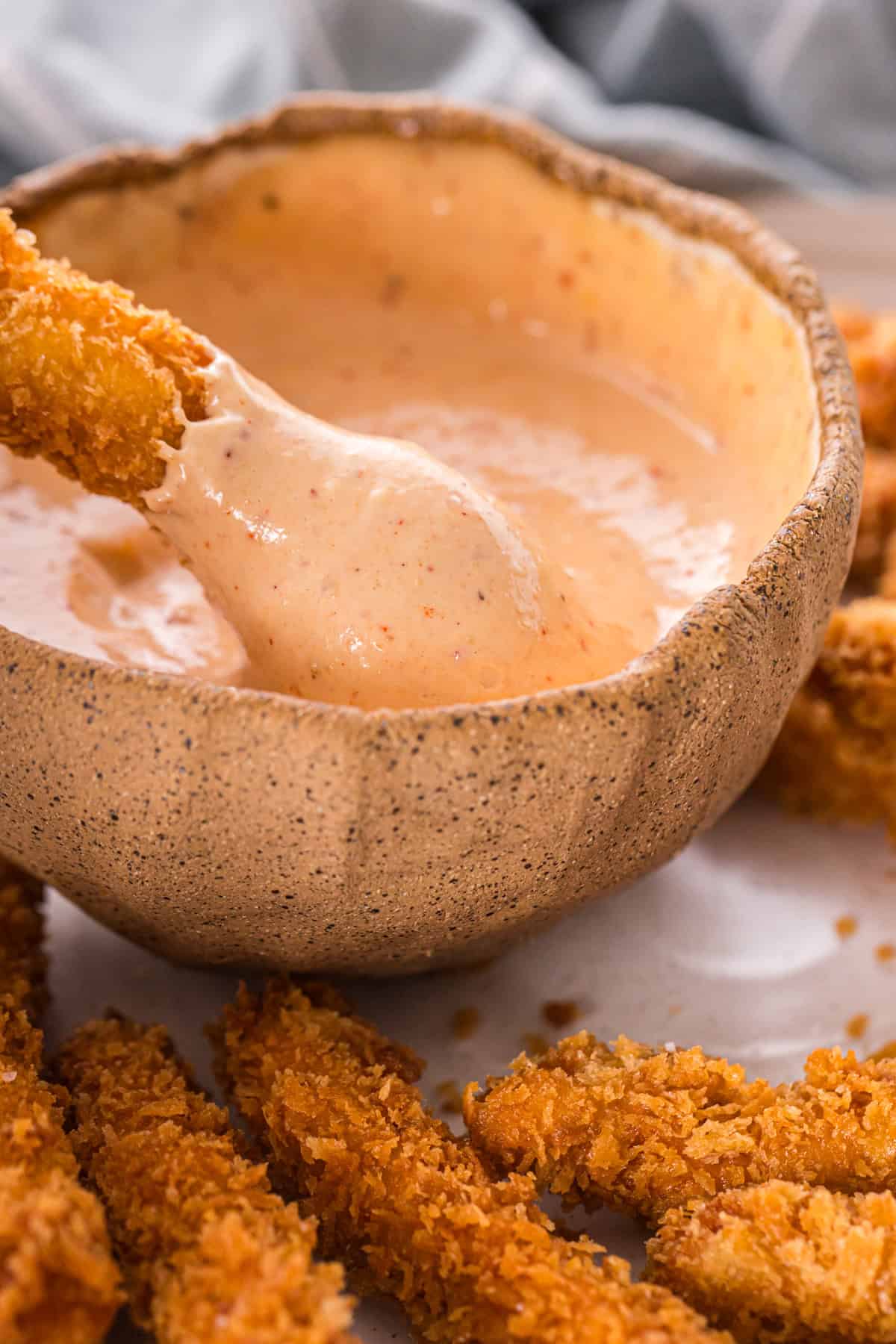 lose-up of a crispy chicken fry dipped into creamy sauce, with more fries in the foreground.