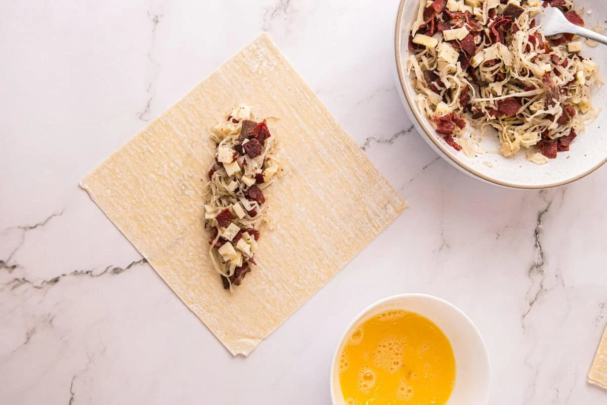 Reuben filling spooned onto the center of an egg roll wrapper, with a bowl of beaten egg for sealing nearby.