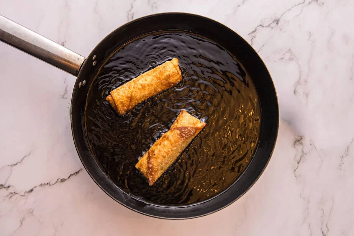 Two Reuben egg rolls frying in a skillet of hot oil until golden brown and crispy.