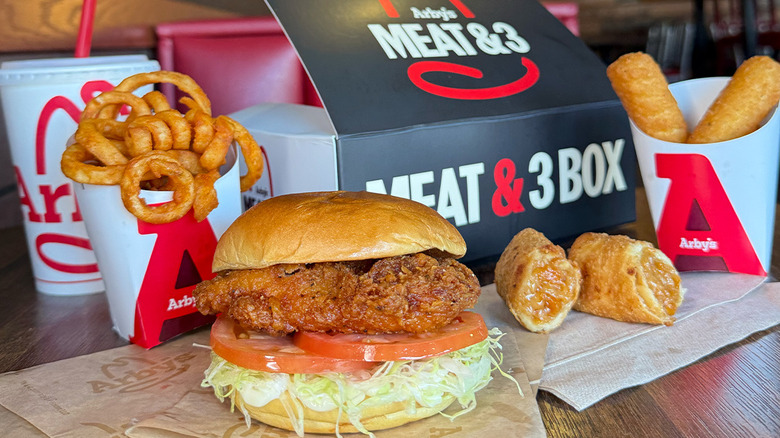 In Arby's chicken sandwich with lettuce and tomato sitting on a table with curly fries, mozzarella sticks, and other Arby's items