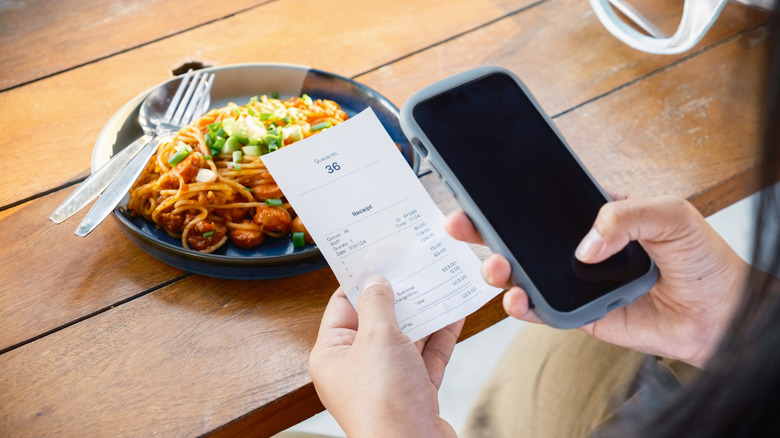 A woman holding a restaurant bill receipt in one hand and her phone in the other over a meal on the table.